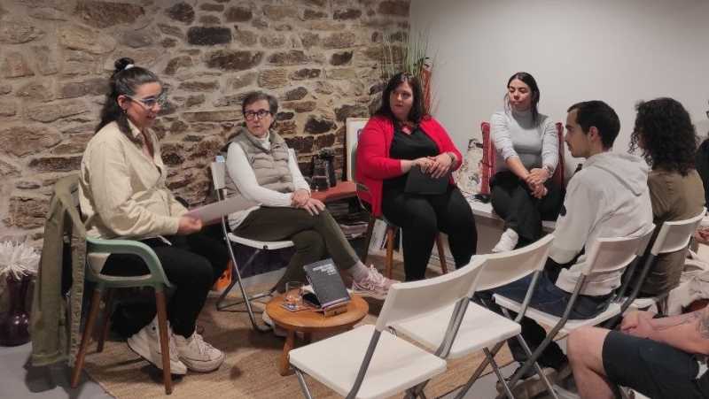 Catro mulleres sentadas cada unha nunha cadeira unha ao lado da outra. Enfronte delas, un home e unha muller tamén sentados e dúas cadeiras baleiras. Entre eles unha mesa baixa de madeira e un libro exposto nela. No fondo hai unha parede de pedra e outra branca facendo esquina. 