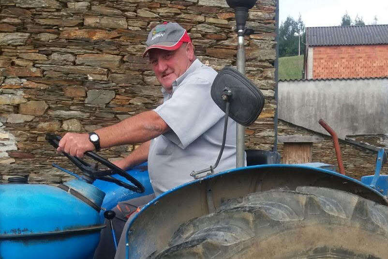 Foto dun home sentado sobre un tractor azul. Leva unha camisa gris, un reloxo negro e unha gorra a xogo con liñas vermellas. Na parte de atrás da foto hai unha casa de pedra e no fondo un cobertizo de cemento xunto a outro de ladrillo.